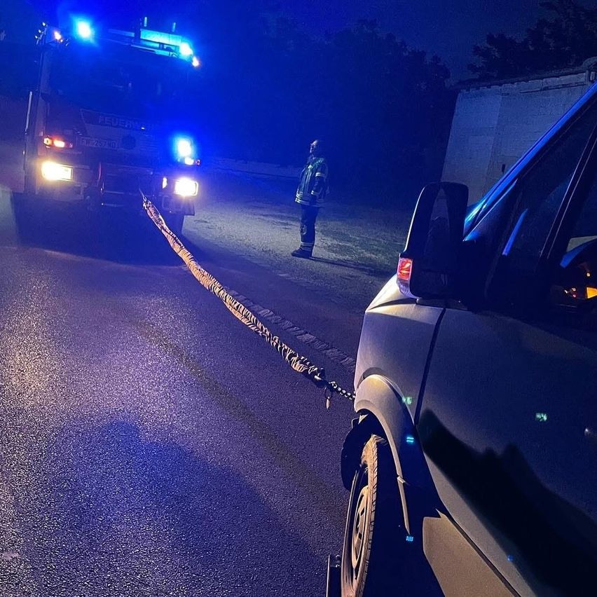 Ein Feuerwehrmann steht im Dunkeln und führt eine am Fahrzeug befestigte Leine vor einem Feuerwehrauto mit blinkenden blauen Lichtern.