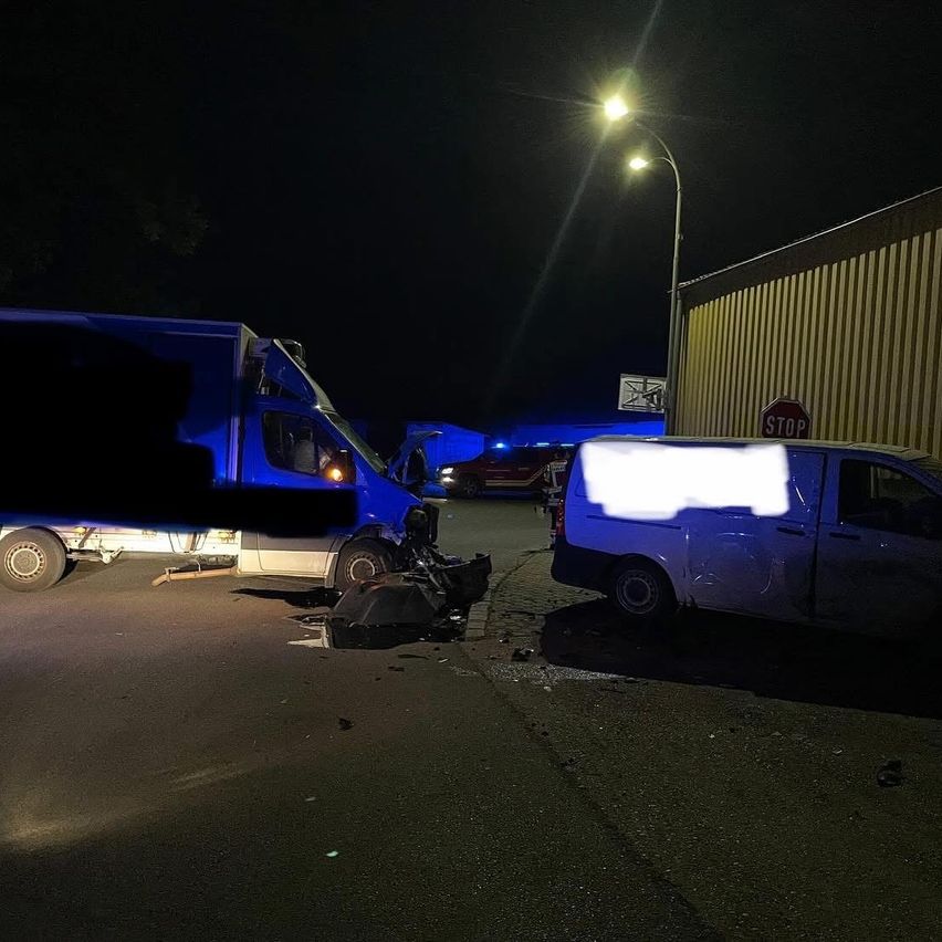 Eine nächtliche Szene einer Straße mit einem blauen und einem weißen Lieferwagen, die verunglückt sind, mit Trümmern und Notleuchten. Ein Stoppschild und eine Straßenlampe sind im Hintergrund zu sehen.