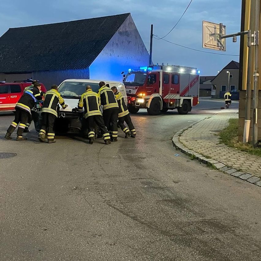 Feuerwehrleute schieben ein Auto am Straßenrand in der Nähe eines Gebäudes und eines Feuerwehrwagens.
