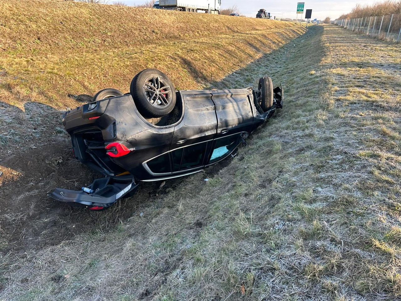 Ein schwarzes Auto ist am Straßenrand umgekippt, mit einem Reifen auf dem Dach und dem anderen auf dem Boden. Das Fahrzeug ist teilweise in Gras vergraben.