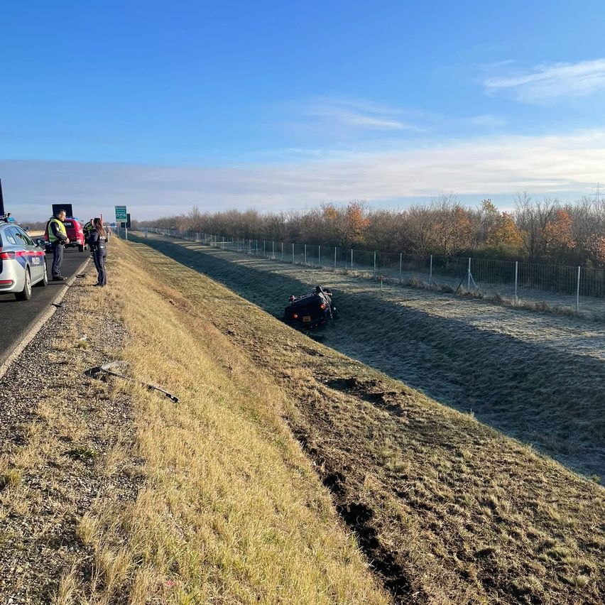Ein Motorrad liegt am Straßenrand, nahe einem grasbewachsenen Hang. Zwei Personen, möglicherweise Polizei, stehen am Straßenrand.