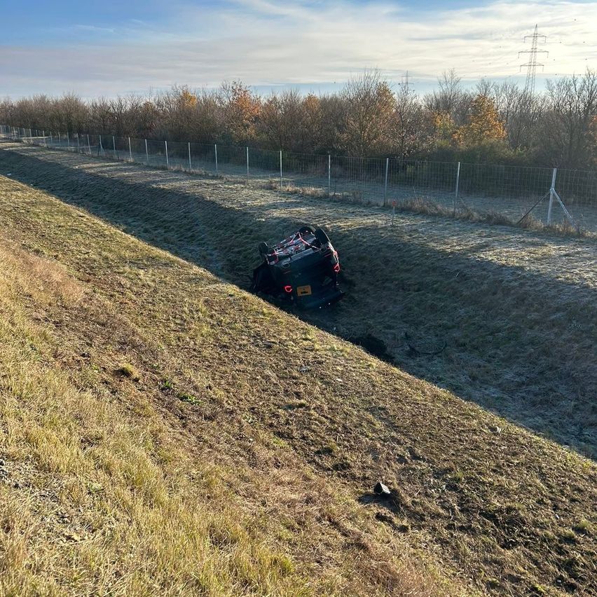 Ein Auto ist in einem Grasfeld in eine Grube gefallen, umgeben von einem Zaun mit Bäumen und Stromleitungen in der Ferne.