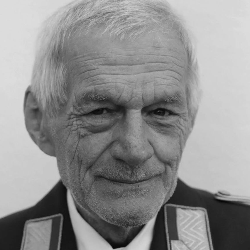 Ein älterer Mann mit weißem Bart und Schnurrbart, in Uniform, schaut direkt in die Kamera und lächelt leicht.