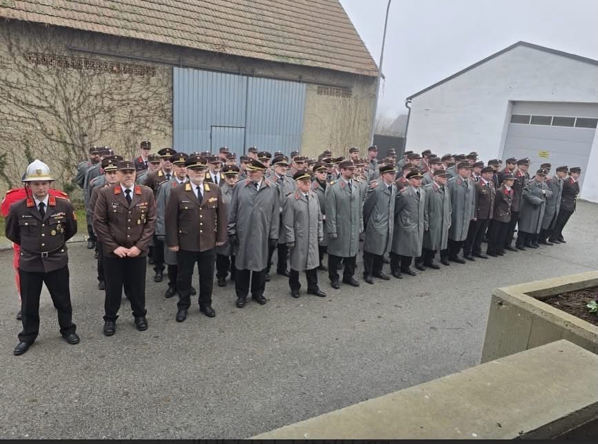 Eine Gruppe von Militärangehörigen in Uniformen und Mützen steht in einer Reihe vor einem Gebäude. Einige tragen Umhänge. Der Boden ist aus Beton.