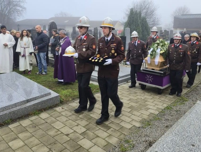 Mehrere Menschen in Uniformen tragen einen Sarg auf einem Friedhof, umgeben von anderen, mit einem Gebäude und Bäumen im Hintergrund.