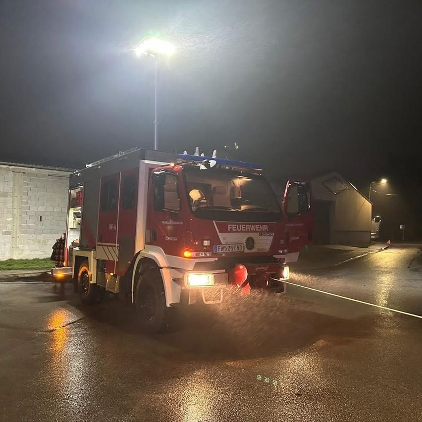 Ein roter Feuerwehrwagen steht nachts auf einer nassen Straße. Die Scheinwerfer des Fahrzeugs sind eingeschaltet und die Tür ist geöffnet.