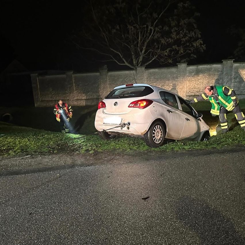 Zwei Feuerwehrleute helfen einem Auto, das nachts von der Straße abgekommen ist. Einer hält eine Taschenlampe und der andere zieht am Auto.