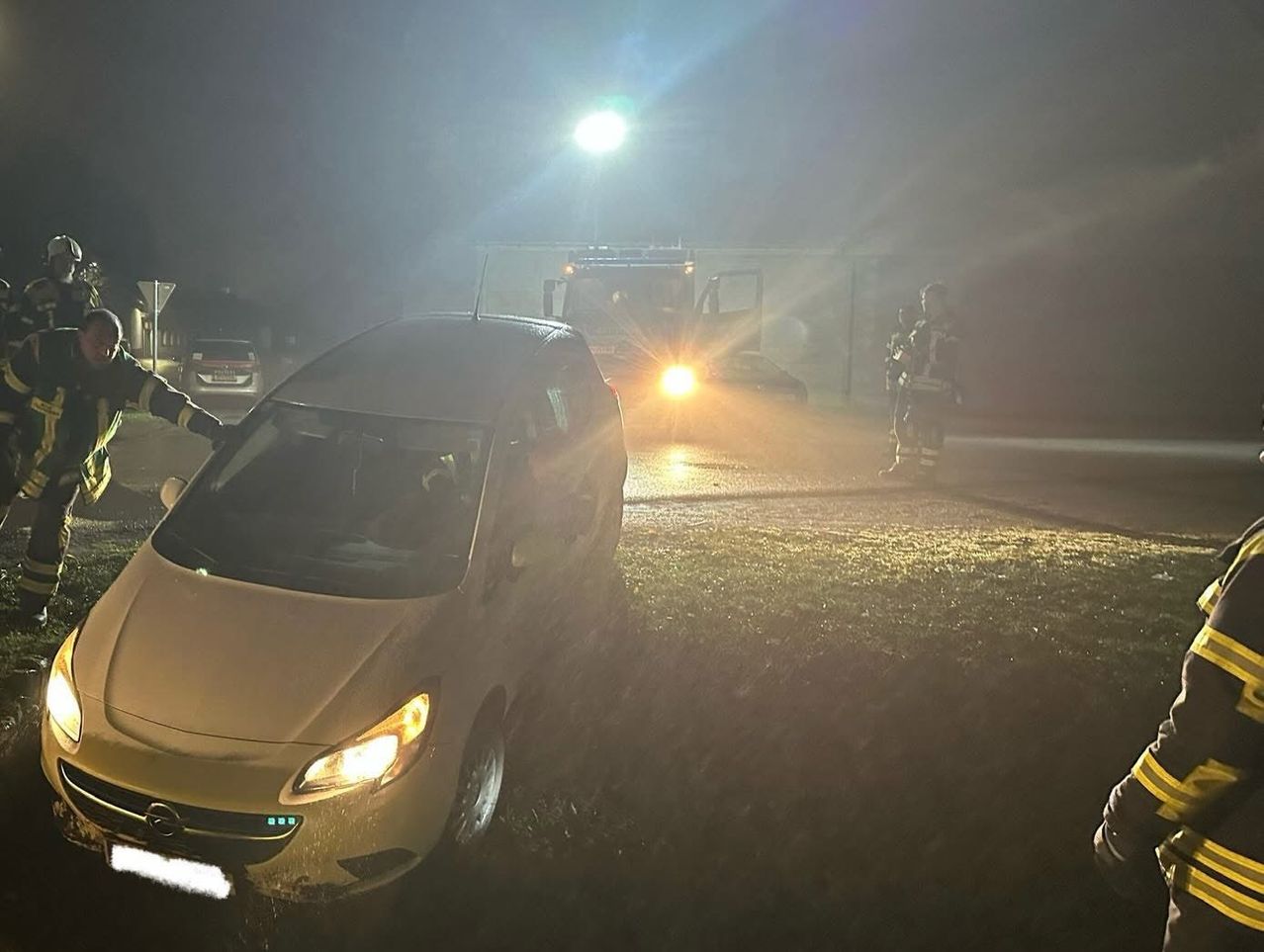 In der Nacht ist ein Fahrzeug auf einem nassen Gelände geparkt, mit Rettungskräften in der Nähe. Ein großes Fahrzeug mit eingeschalteten Scheinwerfern ist ebenfalls anwesend.