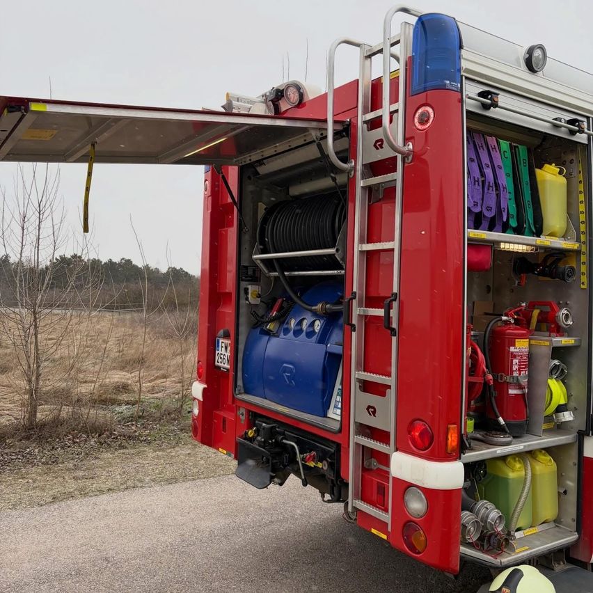 Rückansicht eines roten Feuerwehrwagens mit offener Hecktür, die verschiedene Ausrüstung im Inneren zeigt. Er ist auf einer Straße geparkt.