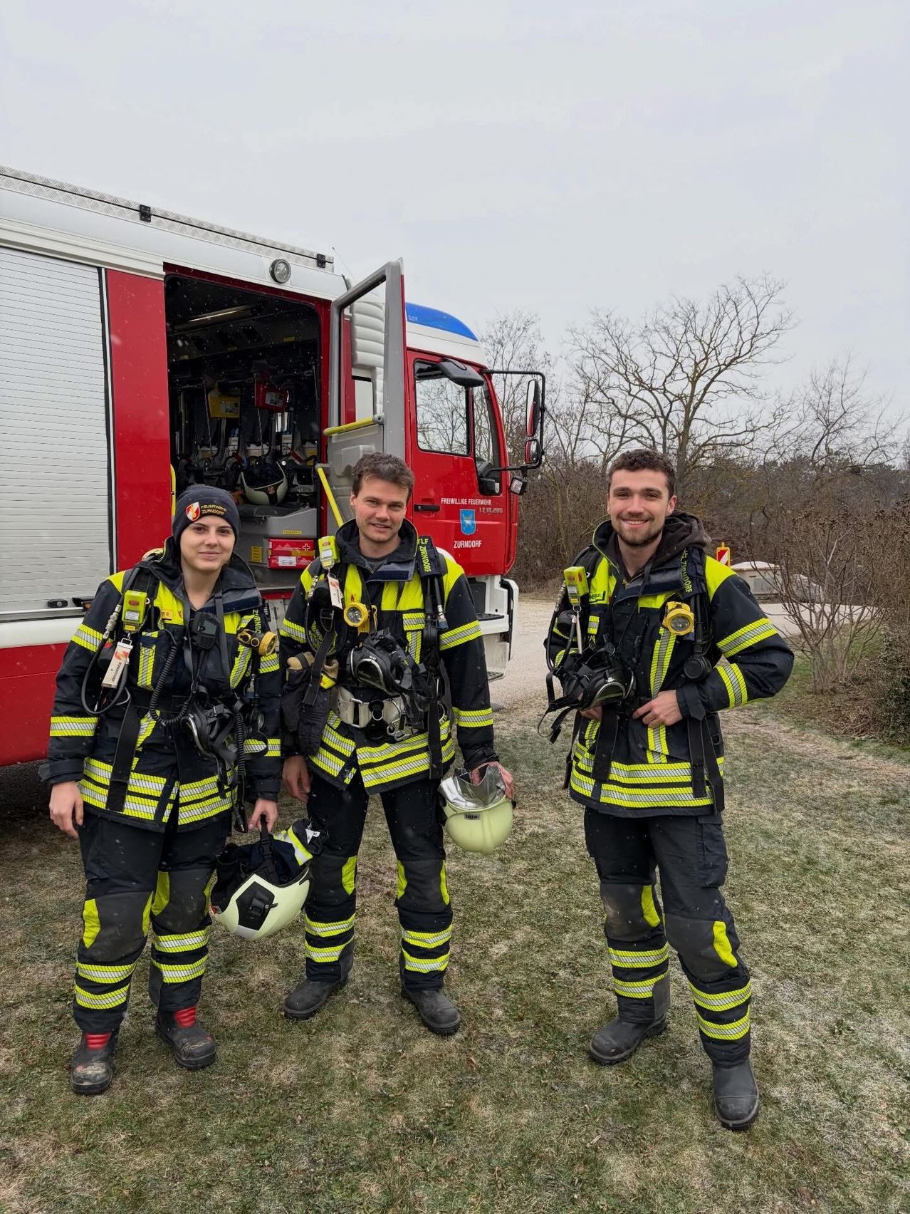 Drei Feuerwehrleute stehen vor einem roten Feuerwehrauto. Sie tragen alle Helme und halten ihre Helme in den Händen.