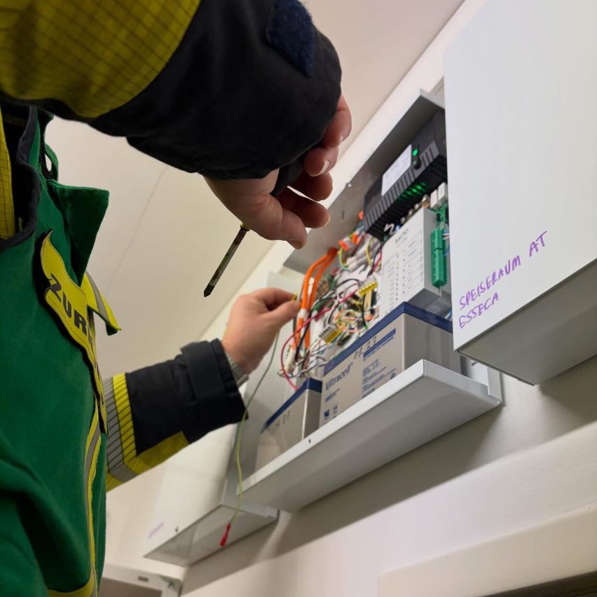 Eine Person in einer grün-gelben Jacke arbeitet an einer elektrischen Schalttafel. Sie benutzt einen Schraubendreher und scheint Anpassungen oder Reparaturen vorzunehmen.