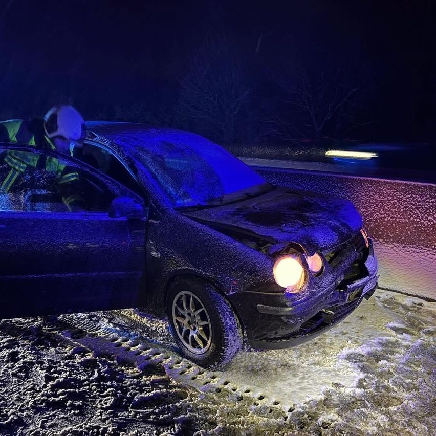 Ein Feuerwehrmann hilft jemandem aus einem blauen Auto mit beschädigter Front auf einer verschneiten Straße bei Nacht.