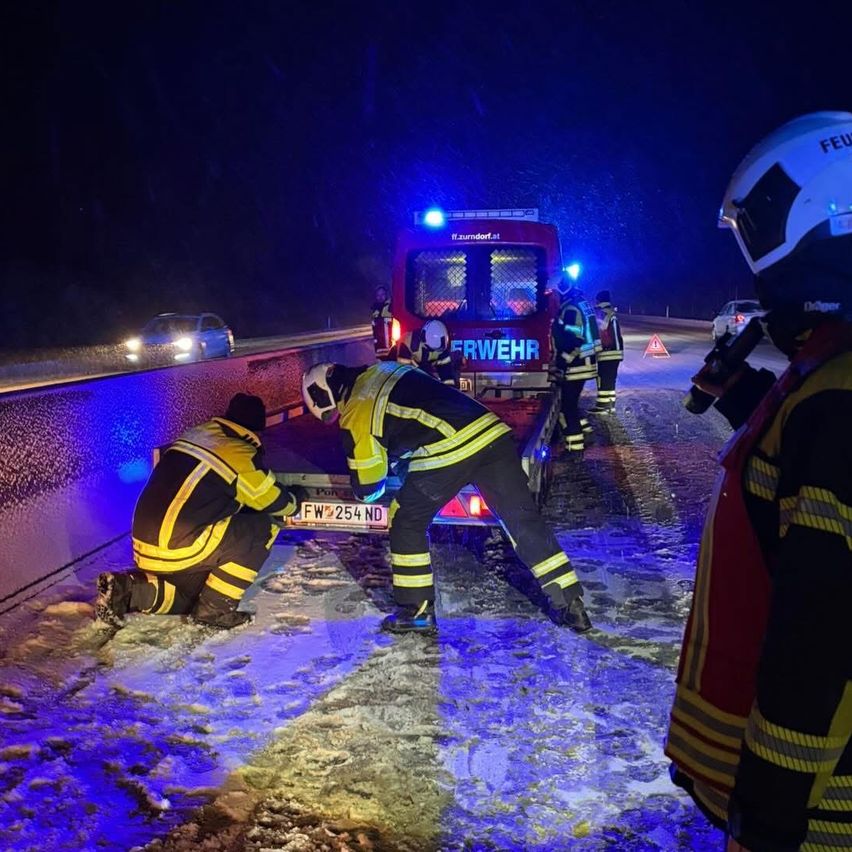 Feuerwehrleute arbeiten an einem schneebedeckten Fahrzeug auf einer Straße bei Nacht. Sie tragen Helme und Uniformen. Ein Feuerwehrwagen mit eingeschalteten Lichtern ist in der Nähe geparkt.