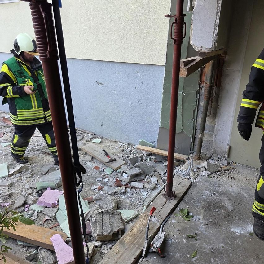 Zwei Feuerwehrleute inspizieren ein beschädigtes Gebäude mit Schutt auf dem Boden und Metallpfosten, die die Struktur stützen. Ein Feuerwehrmann trägt einen Helm und benutzt ein Telefon.
