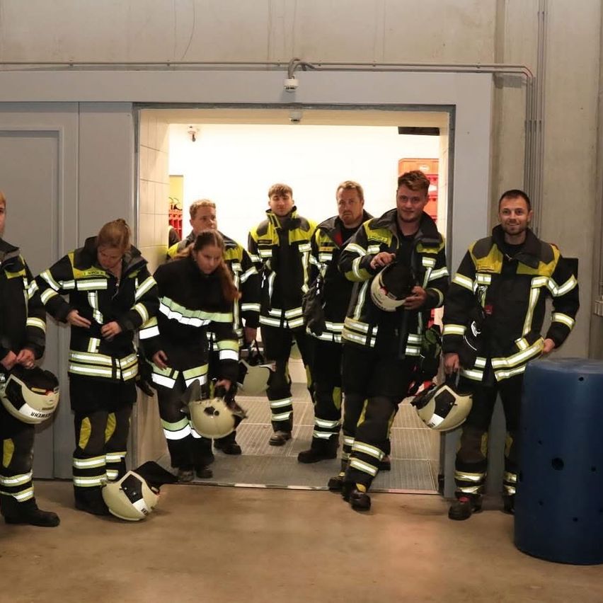 A group of firefighters in full gear stands in front of a door, posing for a photo. Helmets are held in their hands.