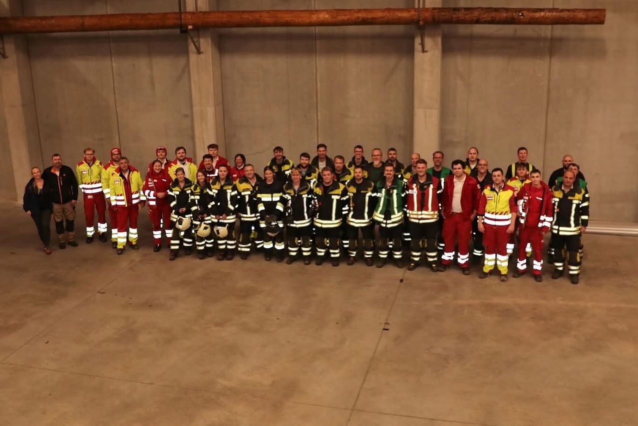 A group of firefighters in safety gear are posing for a photograph inside a building. They are wearing red and yellow uniforms.