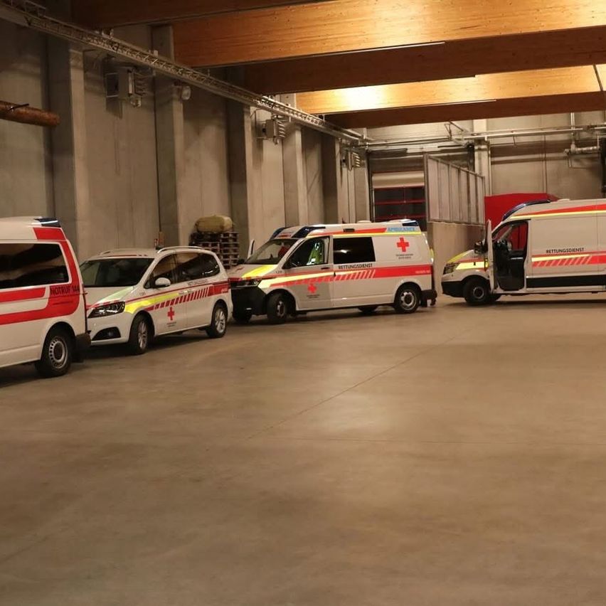 Several ambulances are parked in a warehouse with a concrete floor. The ambulances are white with red stripes and have red crosses on their doors.