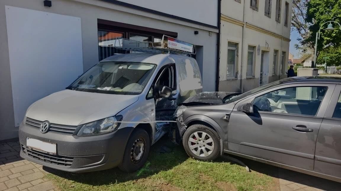 A gray van with a rack on top has collided with a gray car, causing significant damage to the front of the car. The van's door is open.