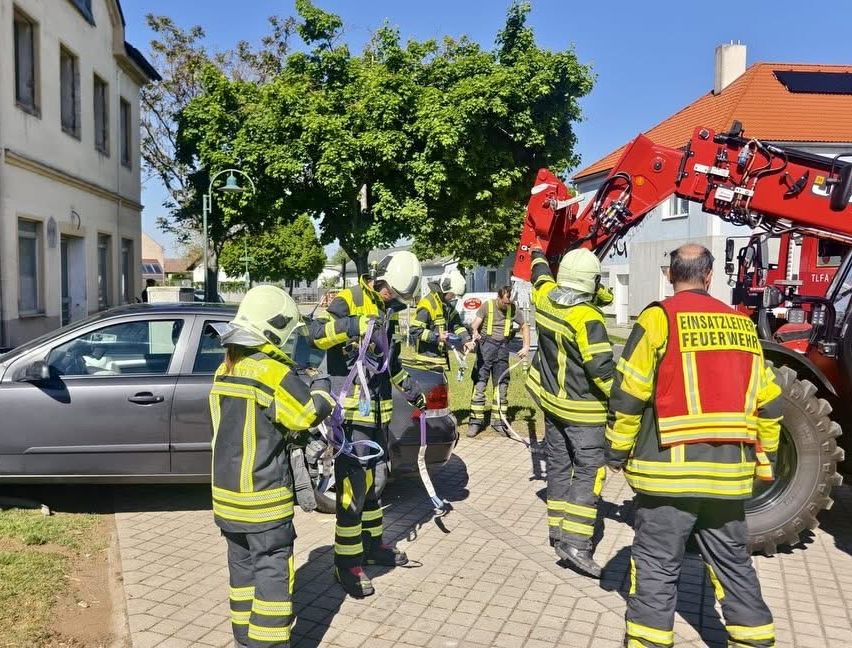 A group of firefighters in full gear are standing on a cobblestone road. A red fire truck is parked nearby, and they are working with ropes. A building and trees are in the background.