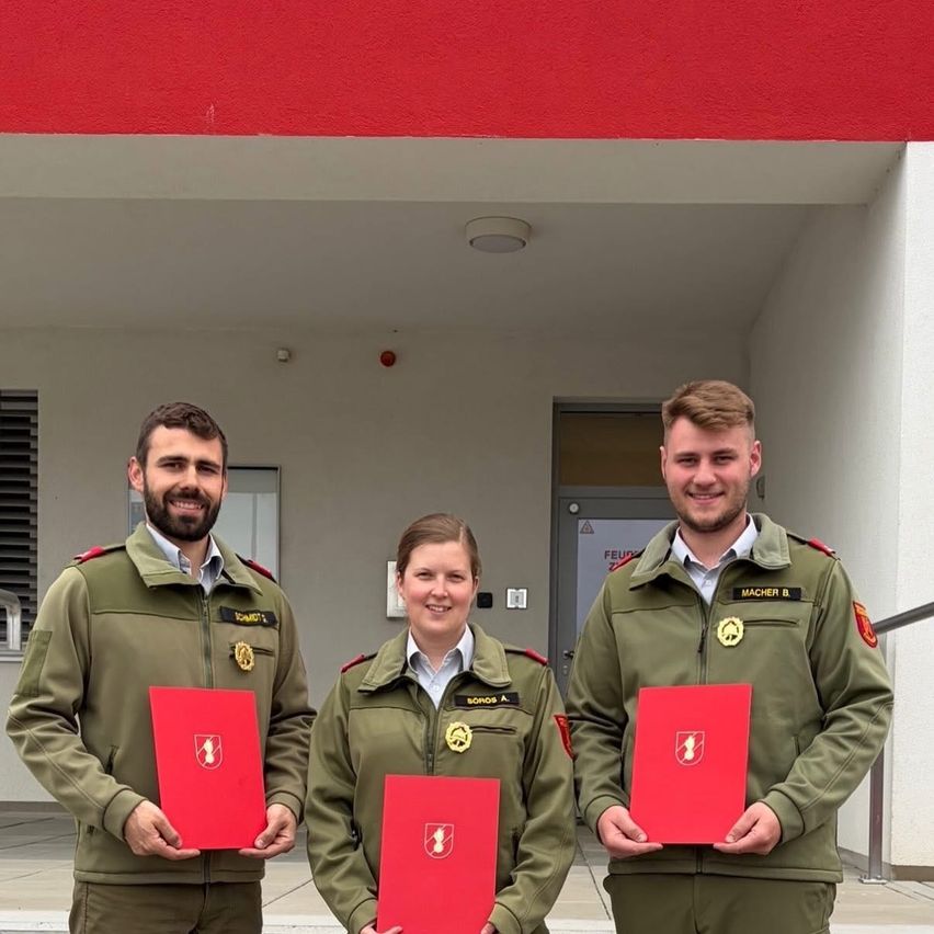 Three individuals, two men and a woman, stand in green uniforms, smiling, in front of a building. They hold red folders with emblems.