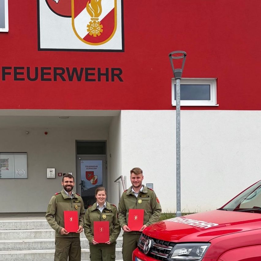 Three individuals in uniform stand outside a red building labeled 'Feuerwehr'. They hold red certificates and are near a red car. A streetlight is visible.