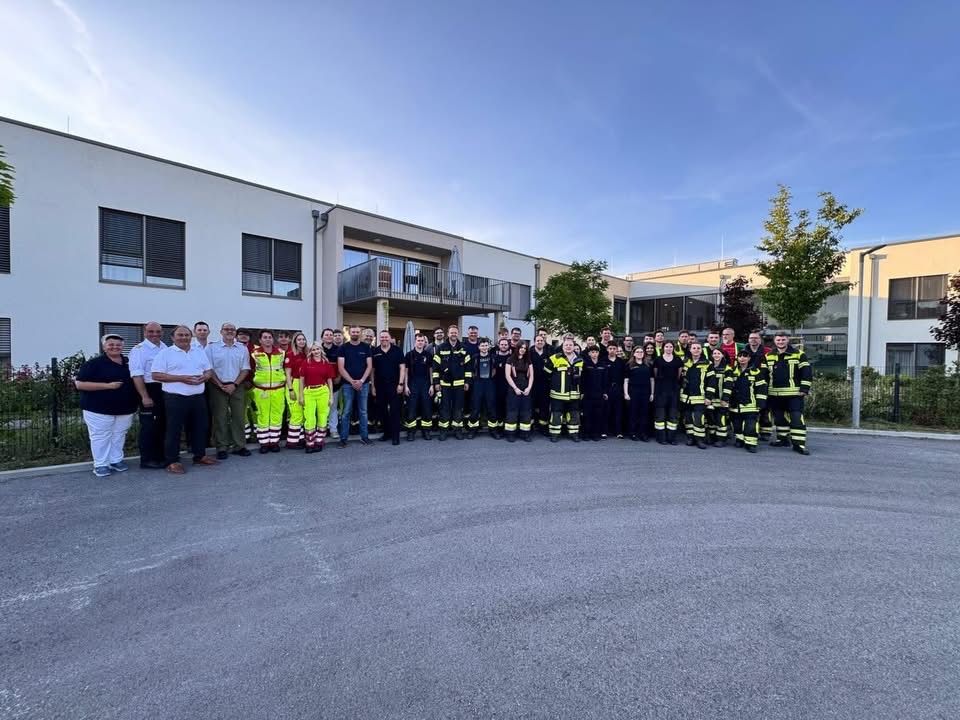 Eine Gruppe von Feuerwehrleuten und Beamten steht vor einem modernen Gebäude bei klarem Himmel. Bäume und Pflanzen sind in der Nähe.
