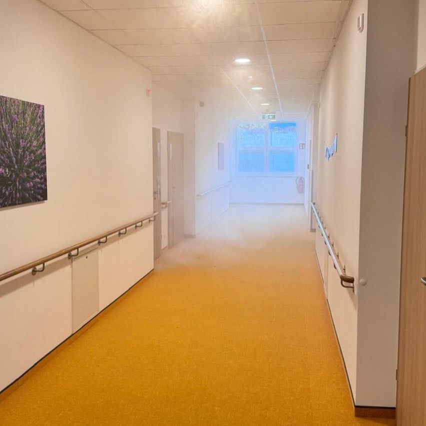 Ein leerer Flur mit gelbem Boden und weißen Wänden. Ein Bild ist an der Wand. Zwei Türen befinden sich auf der linken Seite. Es gibt zwei Handläufe auf beiden Seiten.