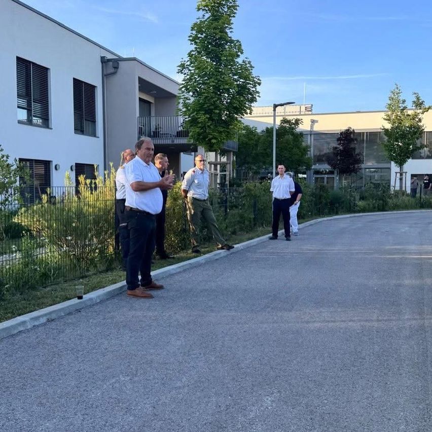 Eine Gruppe von Männern in weißen Hemden steht vor einem Gebäude und bespricht etwas, während andere in der Nähe gehen. Bäume und Pflanzen sind im Hintergrund.