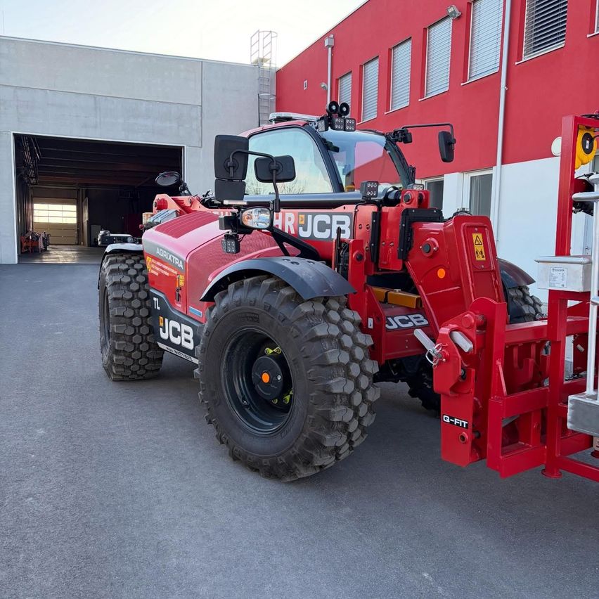 A red JCB tractor is parked in front of a red building. The tractor has large tires and the word JCB on its front.