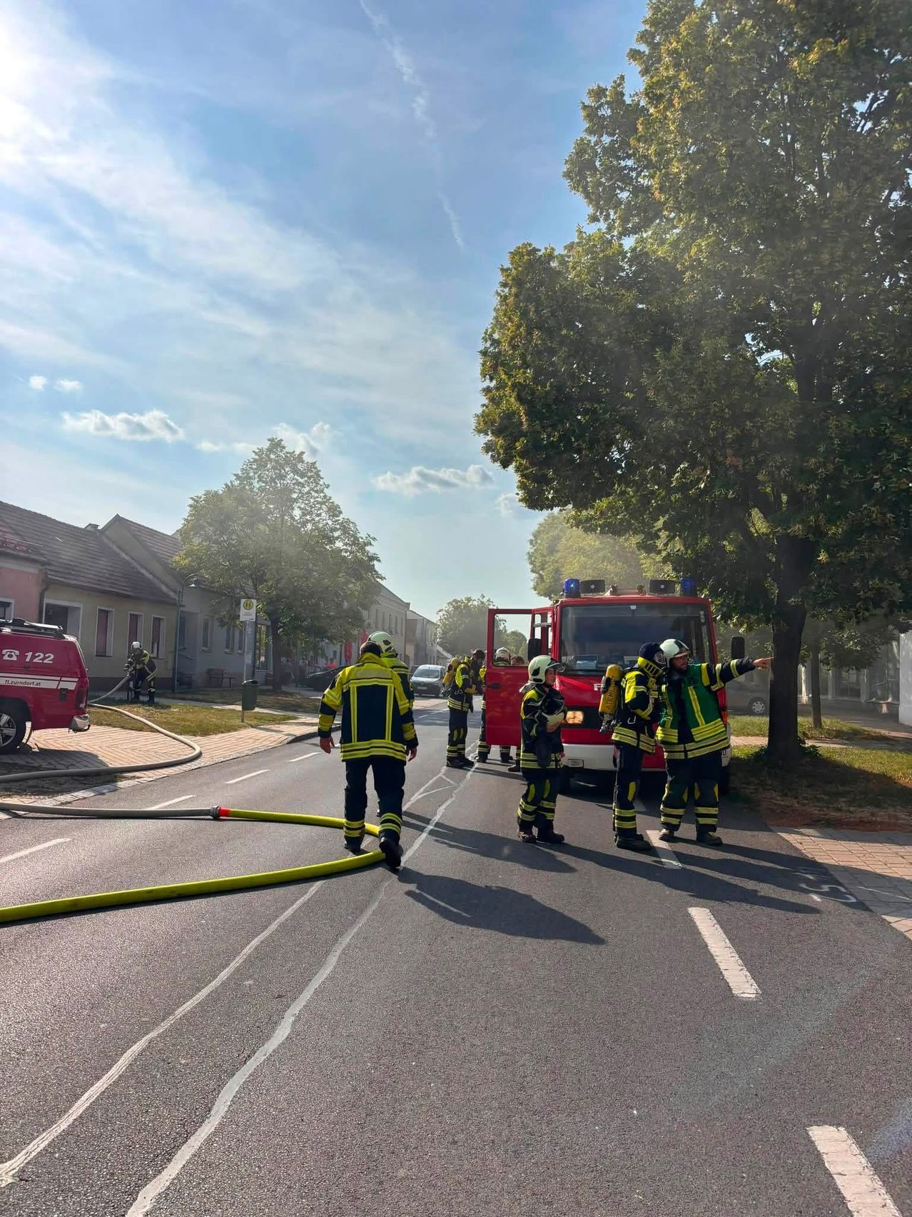 Feuerwehrleute in gelben und schwarzen Uniformen stehen auf einer Straße mit einem roten Feuerwehrwagen im Hintergrund. Ein Feuerhydrant befindet sich auf der linken Straßenseite.
