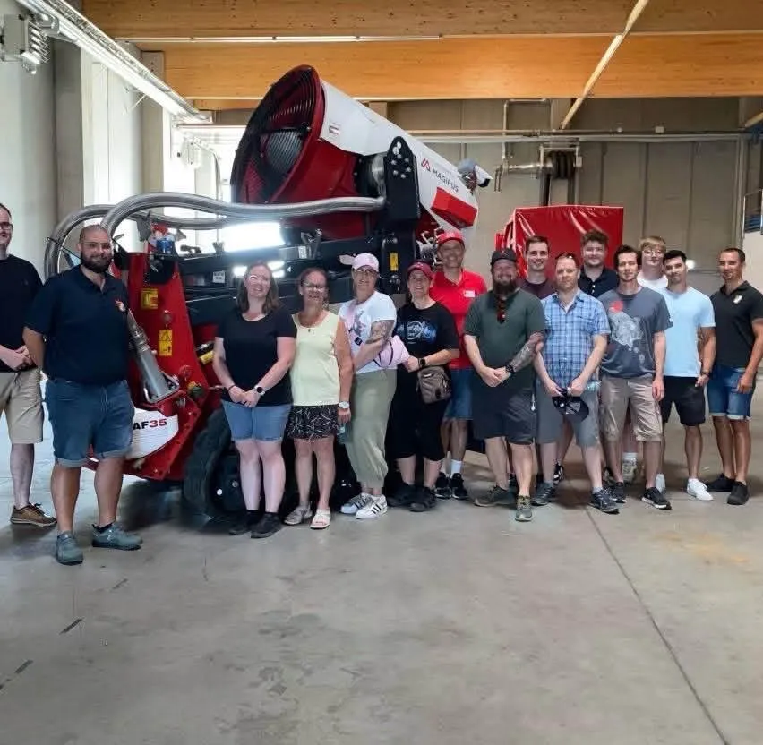 Eine Gruppe von Menschen steht in einem Lagerhaus, lächelt und posiert für ein Foto vor einer großen rot-weißen Maschine.