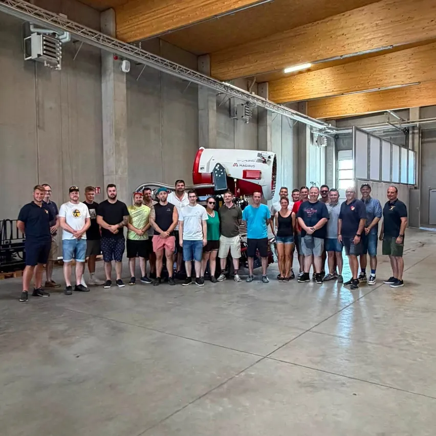 Eine Gruppe von Menschen steht in einer Flugzeughalle mit einem rot-weißen Flugzeug im Hintergrund. Sie sind lässig gekleidet und scheinen für ein Foto zu posieren.