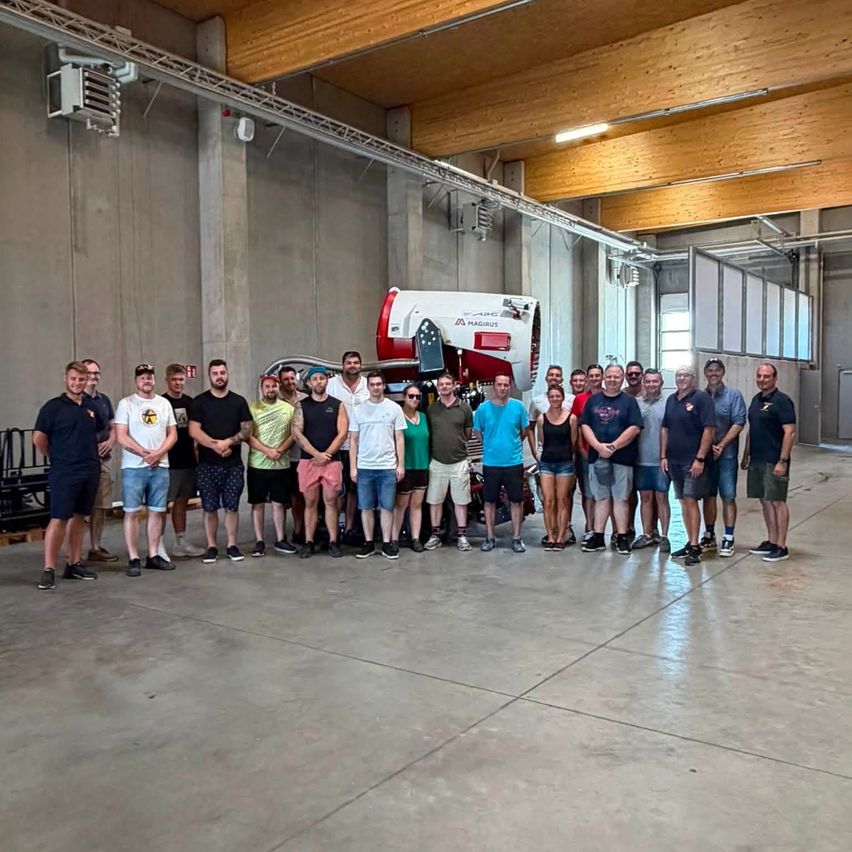 Eine Gruppe von Menschen steht in einer Flugzeughalle mit einem rot-weißen Flugzeug im Hintergrund. Sie sind lässig gekleidet und scheinen für ein Foto zu posieren.