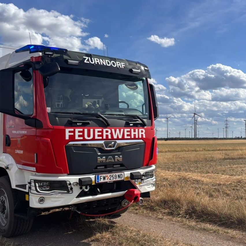 A red MAN fire truck with the word 'Feuerwehr' on the front, parked on a dirt road with a field and wind turbines in the background.