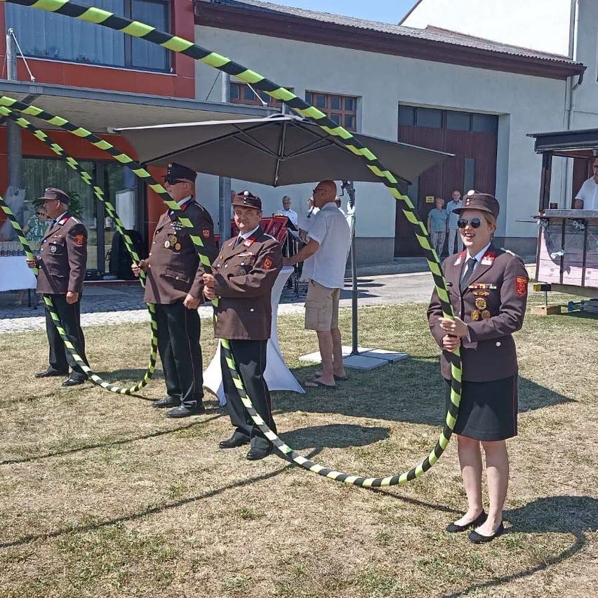 Eine Gruppe von Militärangehörigen in Uniform, die Stäbe halten, steht im Freien. Eine Person trägt eine Sonnenbrille. Sie sind in der Nähe eines Gebäudes mit einem großen Regenschirm.