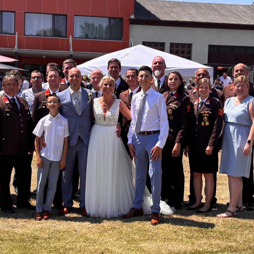 Eine Gruppe von Menschen in formeller Kleidung, einschließlich eines Brautpaares, posiert für ein Foto auf einem Rasen. Sie stehen vor einem roten Gebäude und einem weißen Zelt.
