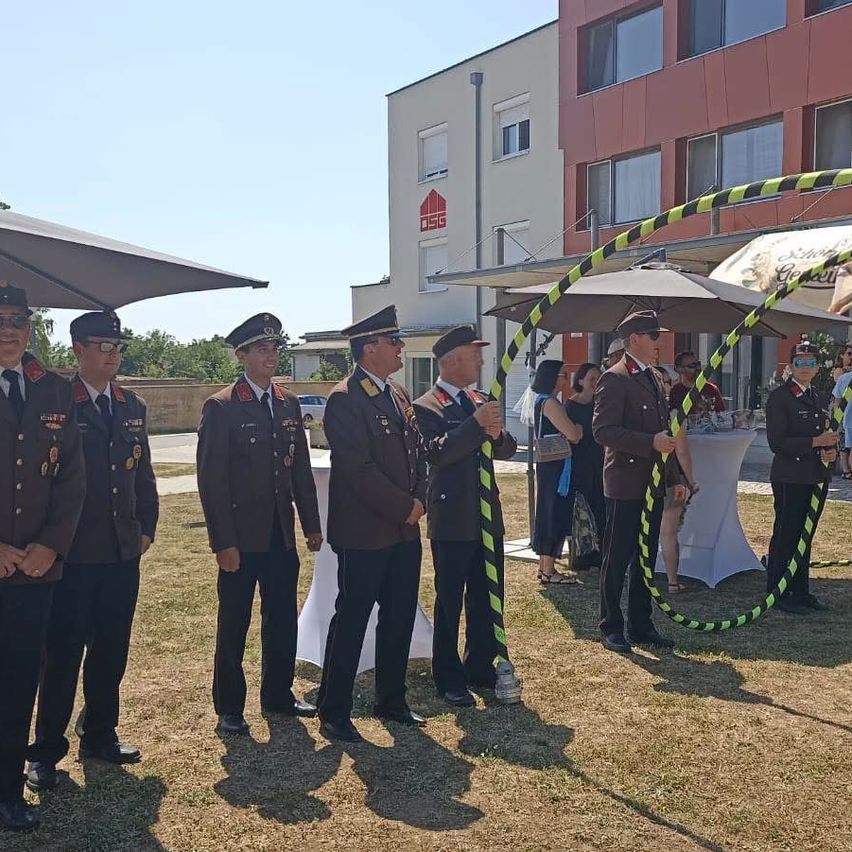 Fünf uniformierte Männer stehen mit einem großen Hula-Hoop draußen. Ein großes Gebäude und ein Baldachin sind hinter ihnen. Eine Gruppe von Menschen steht in der Nähe, einige unter einem Regenschirm.