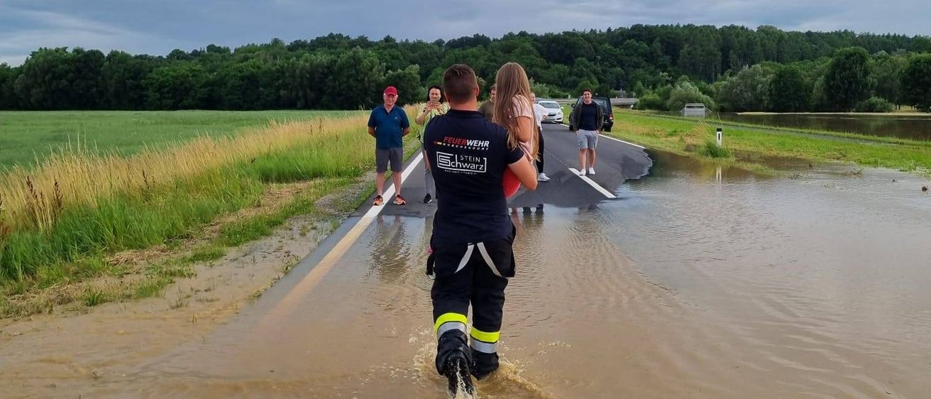 Ein Rettungshelfer watet durch das Hochwasser und trägt eine Frau in Sicherheit. In der Nähe stehen ein Mann und eine Frau auf der Straße, ein Auto parkt in der Ferne.