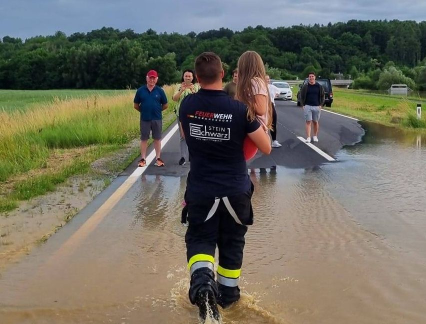 Ein Rettungshelfer watet durch das Hochwasser und trägt eine Frau in Sicherheit. In der Nähe stehen ein Mann und eine Frau auf der Straße, ein Auto parkt in der Ferne.