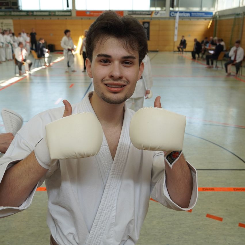 Ein Mann in einem weißen Karateanzug hält gepolsterte Handschuhe in einer Sporthalle hoch und lächelt. Im Hintergrund sitzen und stehen Personen.