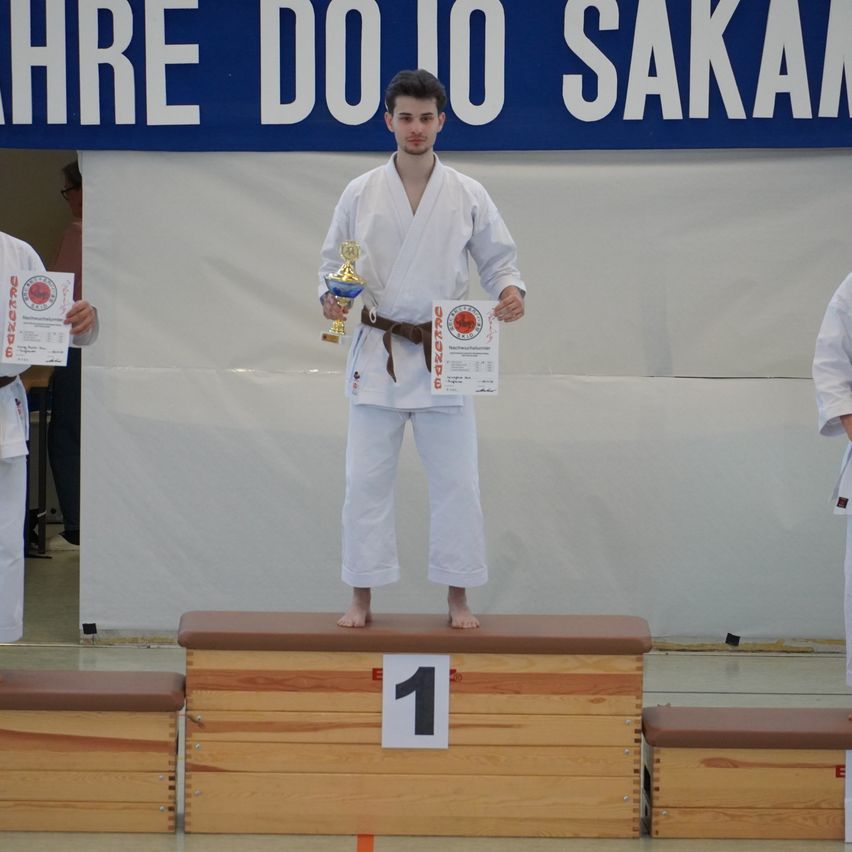 Ein Mann steht auf einem hölzernen Podest und feiert mit einer Trophäe und einem Zertifikat. Dahinter hält eine andere Person ein Zertifikat. Ein blauer Banner steht 'AHRE DO IO SAKAN'.