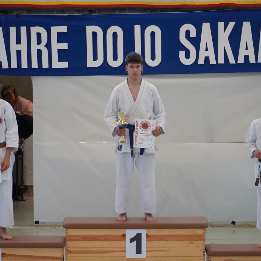 Ein junger Mann steht auf einem Podest mit einer Medaille, hält eine Trophäe und ein Zertifikat. Er trägt ein weißes Karate-Uniform. Hinter ihm steht ein Banner mit der Aufschrift 'Jahre Dojo Saka'. Zwei weitere Personen in weißen Uniformen stehen an den Seiten.