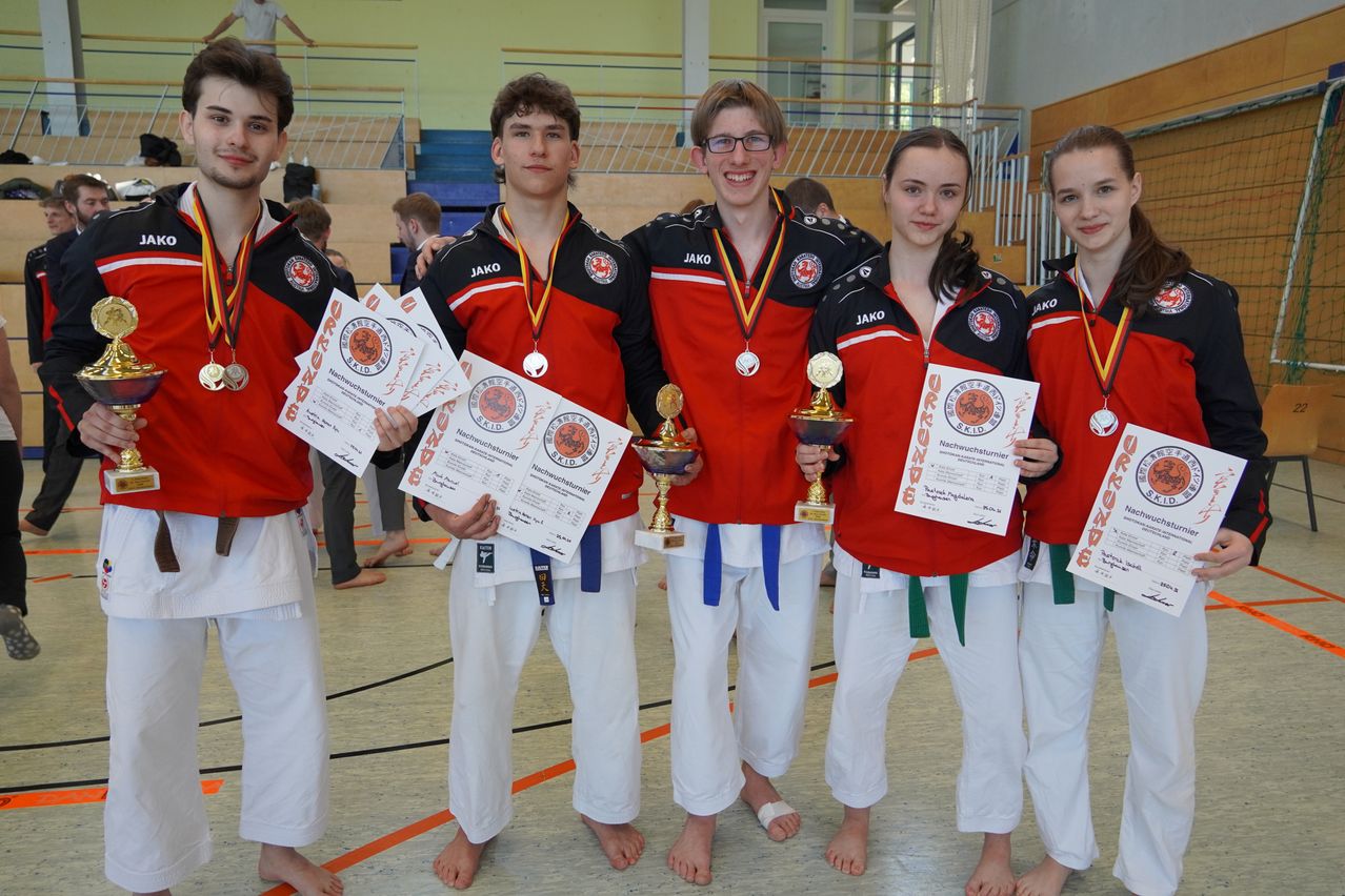 Fünf Personen in roten und weißen Kampfsportuniformen stehen zusammen und halten Medaillen und Pokale. Sie scheinen einen Sieg bei einem Wettbewerb zu feiern.