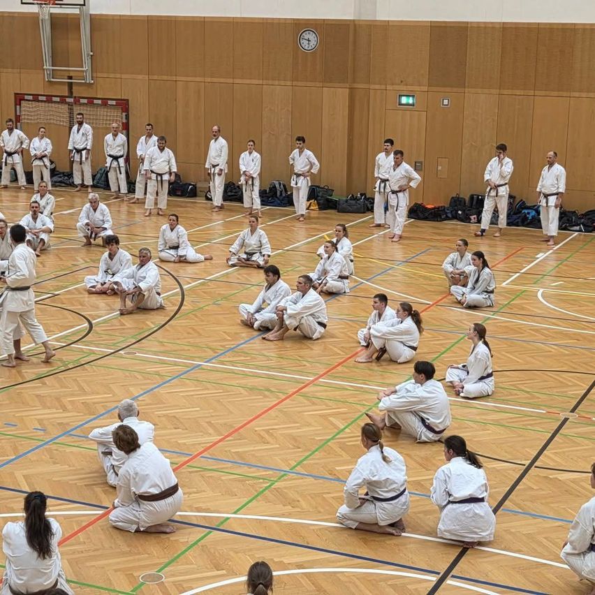 A group of people in white uniforms are practicing karate in a gym. Some are seated while others are standing.