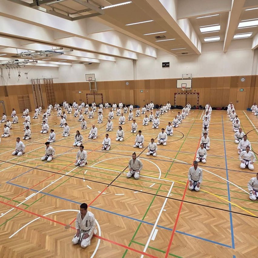 A group of people in white uniforms are practicing martial arts in a large indoor gym. They are sitting in a circle on the wooden floor. There are basketball hoops on the side.