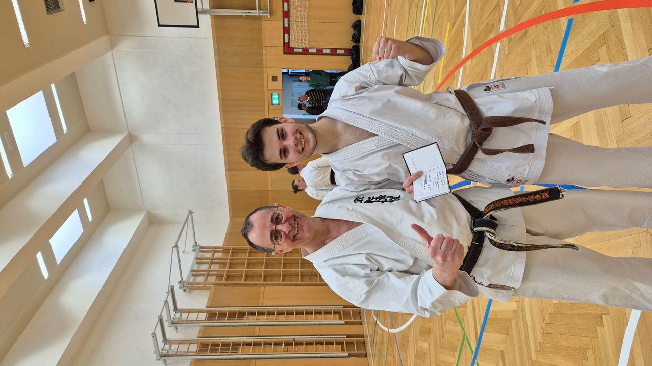 Two men in white judo uniforms are smiling and posing for a photo. One holds a certificate, and they are in a gym.