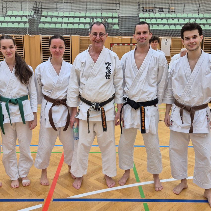 Five people in white martial arts uniforms stand together on a court, with two women on the left, two men in the middle, and a young man on the right. Behind them is a wooden barrier with empty green chairs.