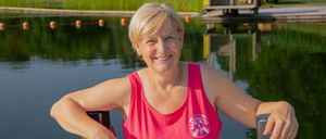 Eine Frau in einem rosa Tanktop steht am Wasser, lächelt und lehnt sich an einen Holzzaun. Hinter ihr erstreckt sich ein Steg in den See, mit Bojen, die im Wasser schwimmen.