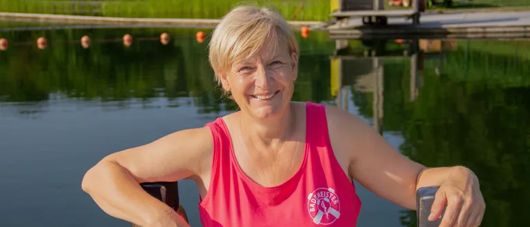 Eine Frau in einem rosa Tanktop steht am Wasser, lächelt und lehnt sich an einen Holzzaun. Hinter ihr erstreckt sich ein Steg in den See, mit Bojen, die im Wasser schwimmen.