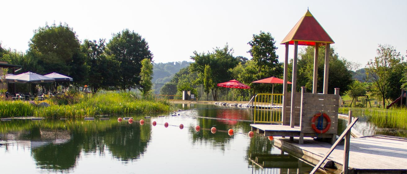 Ein ruhiger Wasserpark mit einem kleinen Spielplatz, umgeben von üppiger Grünfläche und Bäumen. Das Wasser ist ruhig, mit roten und weißen Bojen, die einen sicheren Schwimmbereich markieren.